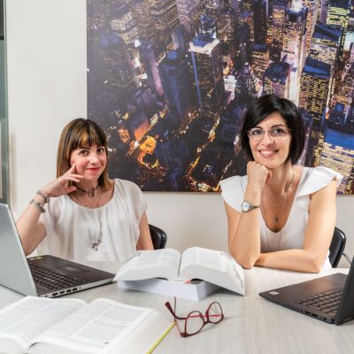 Avvocati Paola Gallozzi e Alessandra Fischetti che indossano una maglietta bianca. Sopra la scrivania vi sono due computer portatili con dei libri di legge